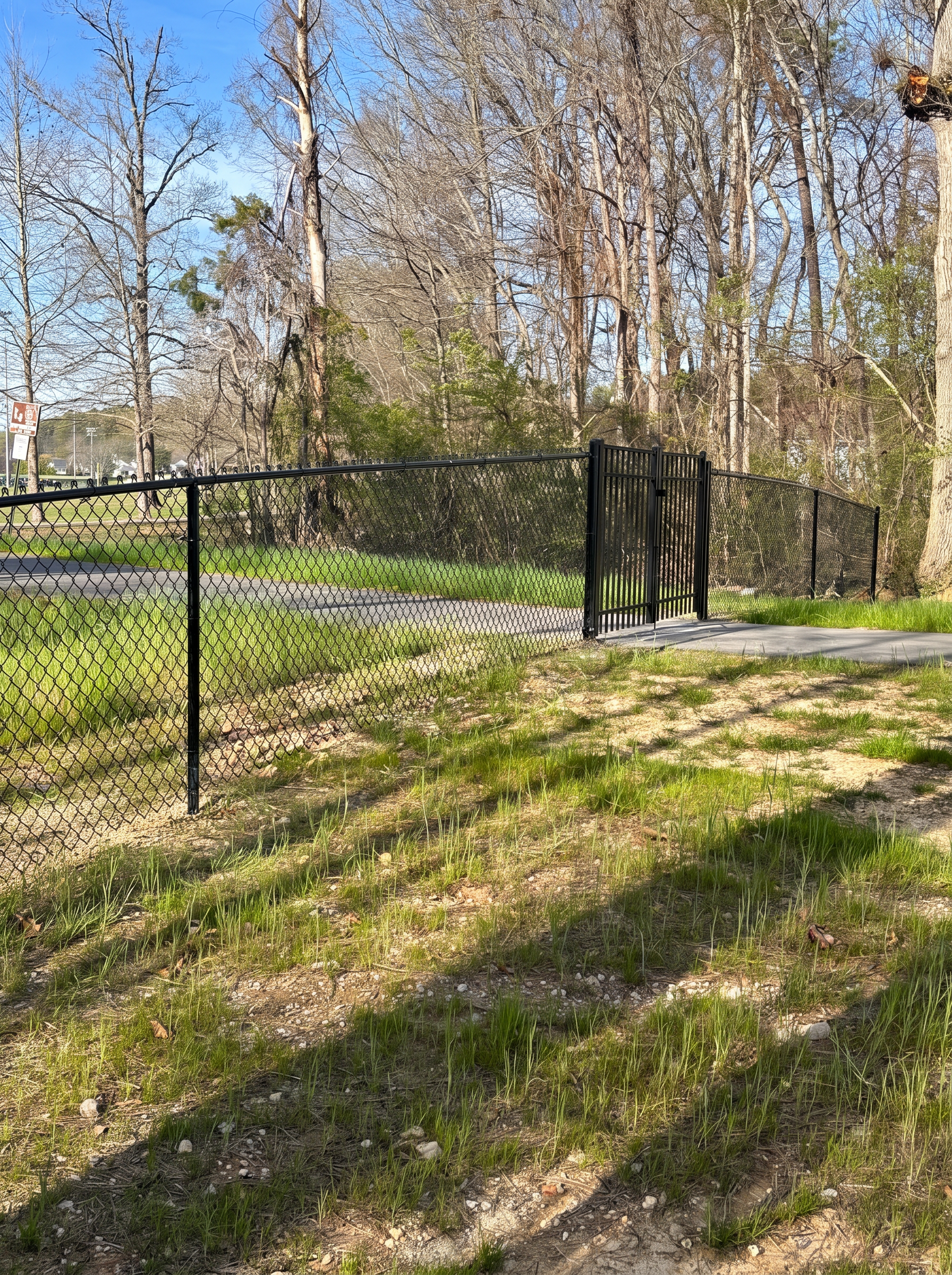 Black chain-link perimeter fence with an ornamental walk gate on a wooded paved path