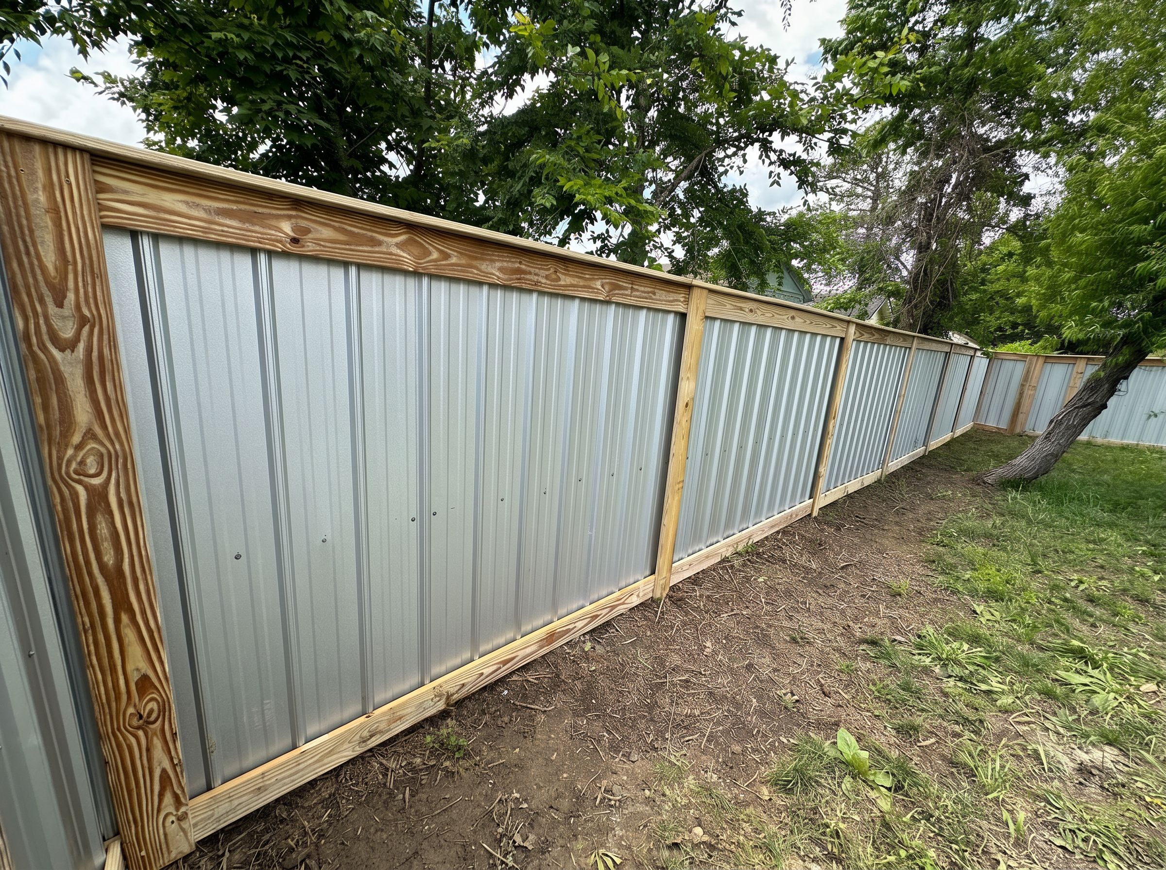 Long corrugated aluminum privacy fence with wood posts and cap beneath shade trees